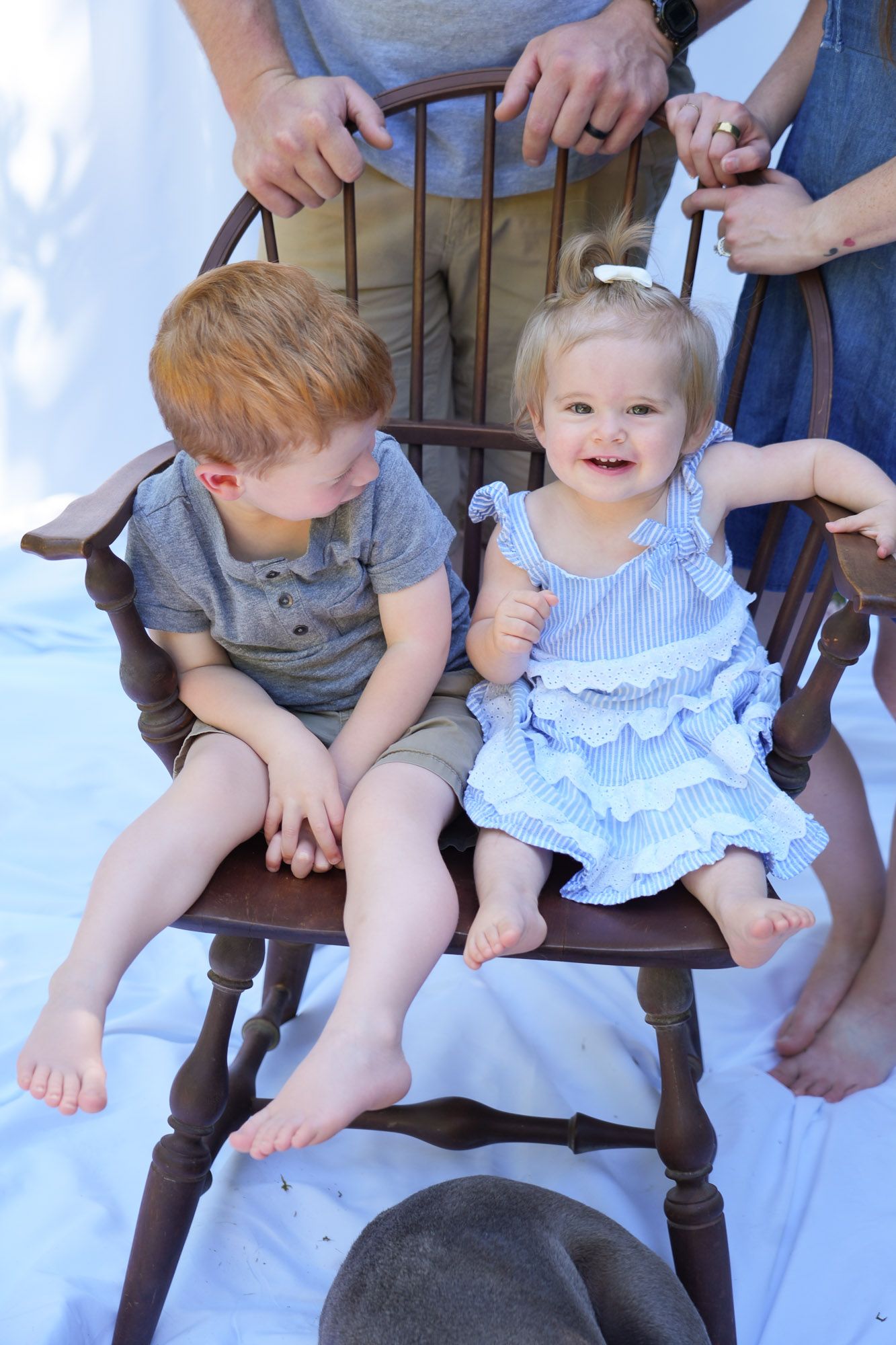 Red-haired boy and smiling blonde toddler girl share wooden chair during outdoor family portrait session.