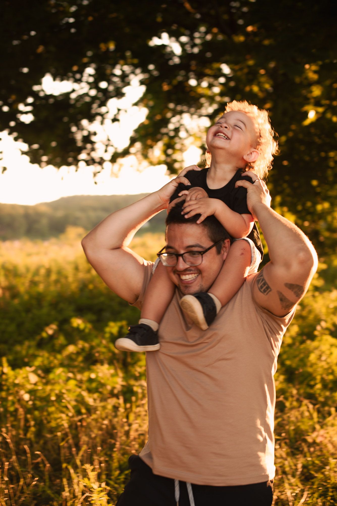 A laughing toddler sits on his smiling father's shoulders in a sunny green field at sunset.