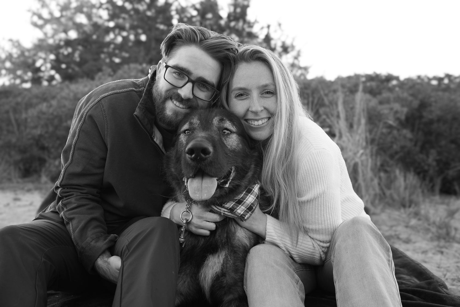 Black and white photo of couple posing with their German Shepherd dog on a beach.