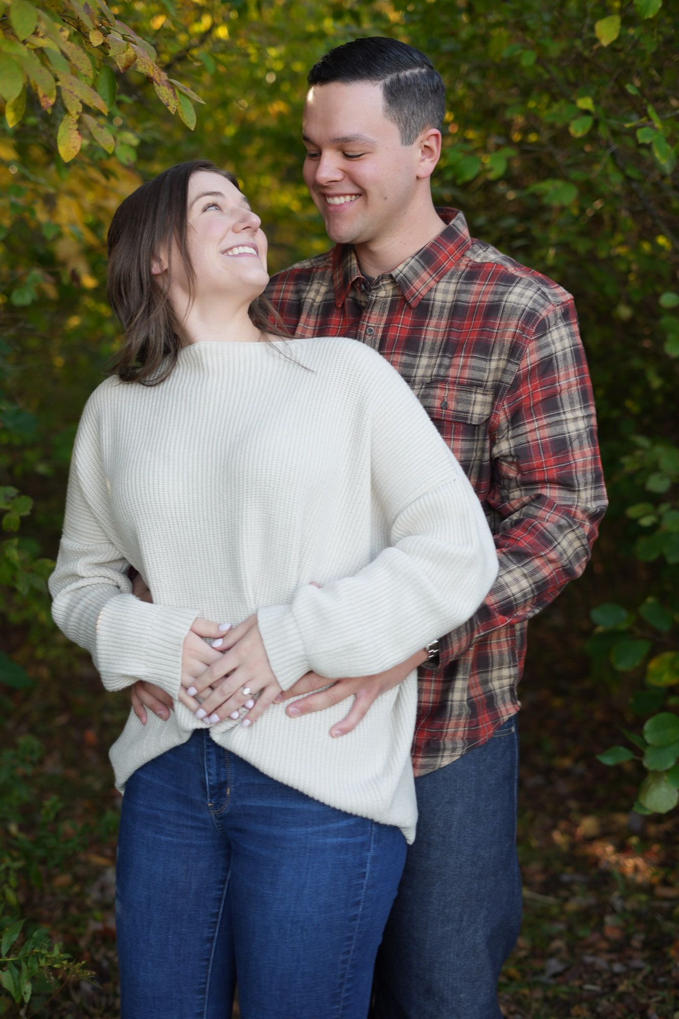 Laughing couple embracing outdoors with autumn foliage, woman in cream sweater and man in plaid shirt.
