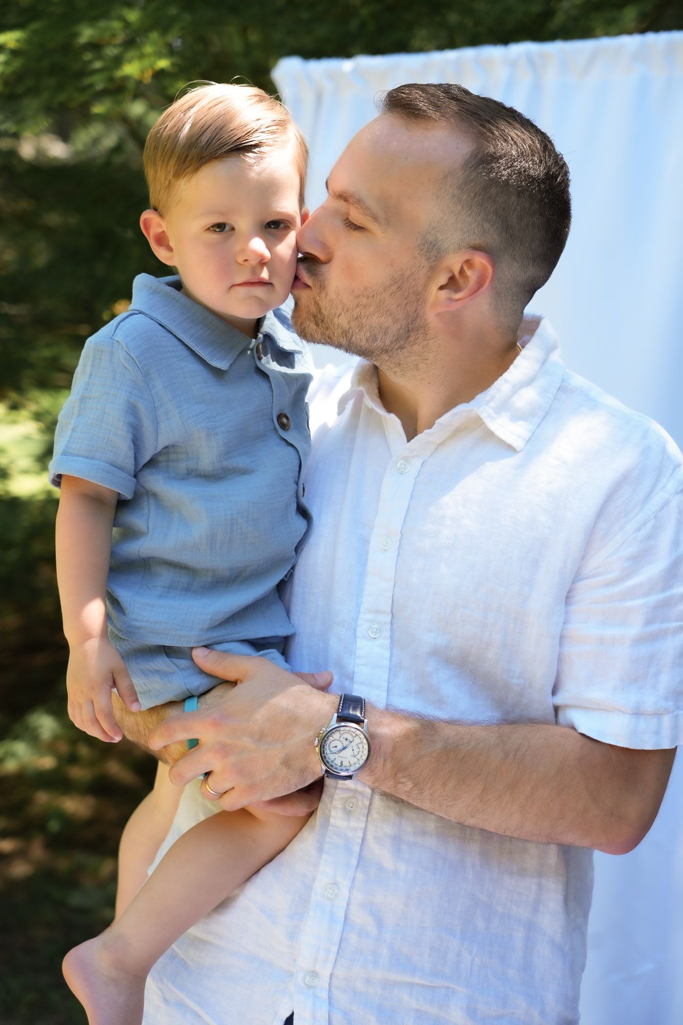 Father kissing toddler boy on cheek outdoors during family photo session.