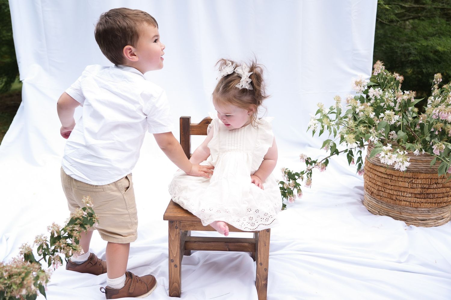 Young boy in white shirt interacts with smiling toddler girl in white dress seated on small wooden chair outdoors.
