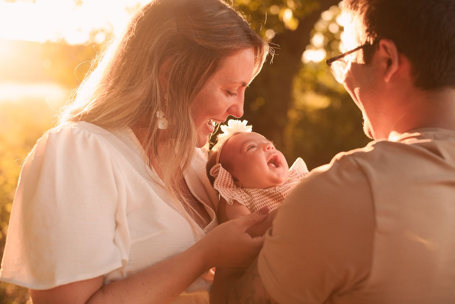 A mother cradles her laughing newborn while a father looks on lovingly in golden sunlight.