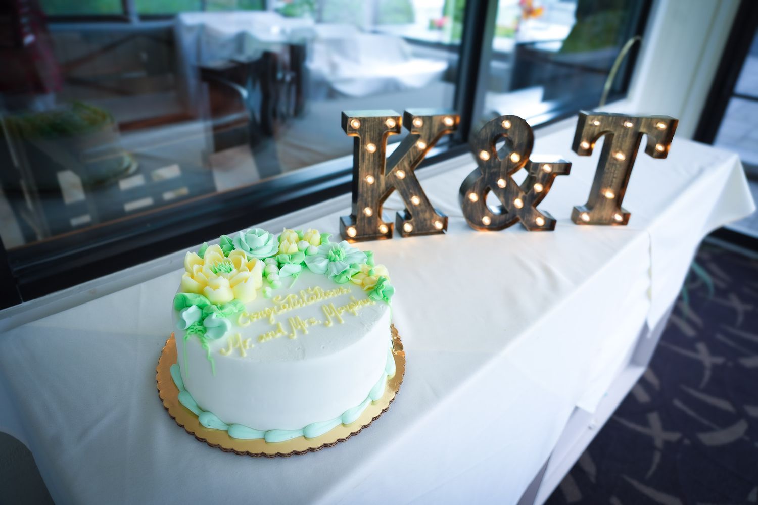 Elegant white wedding cake with green floral accents displayed beside glowing K&T marquee letters.