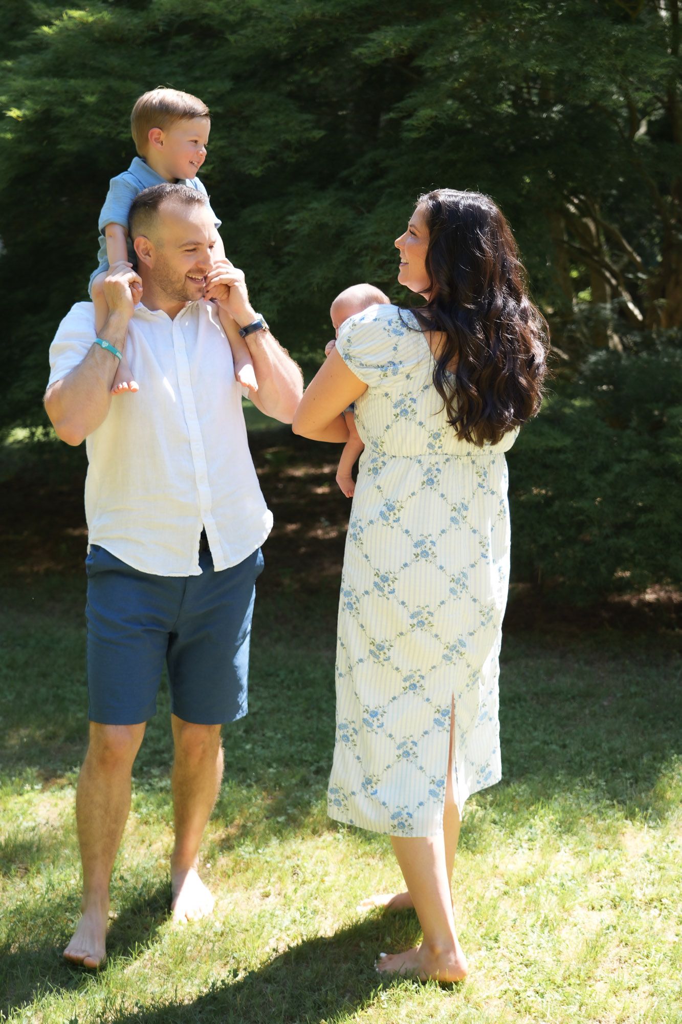 Young family walking barefoot on grass, dad with toddler on shoulders, mom holding newborn.