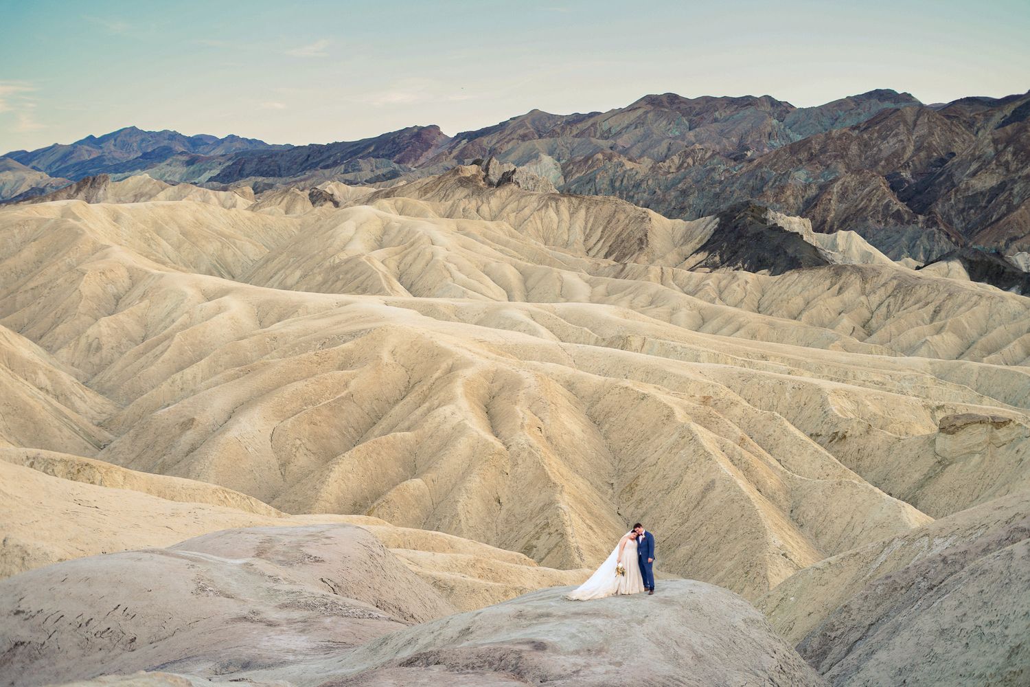 Bride and groom pose in white dress and blue suit amid dramatic Death Valley badlands terrain at Zabriskie Point.