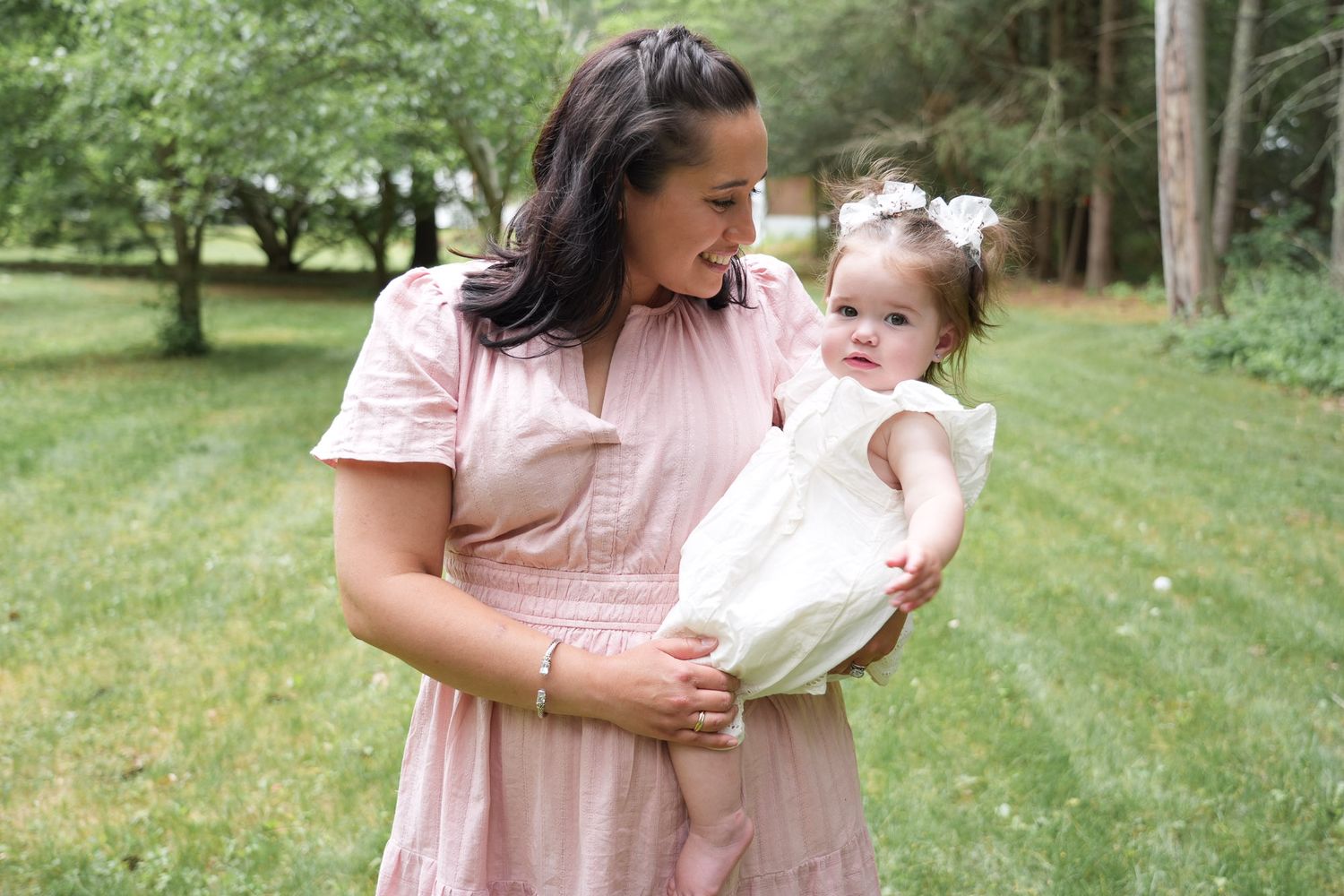 A mother in a pink dress tenderly holds her baby daughter in a white dress with a bow headband outdoors.