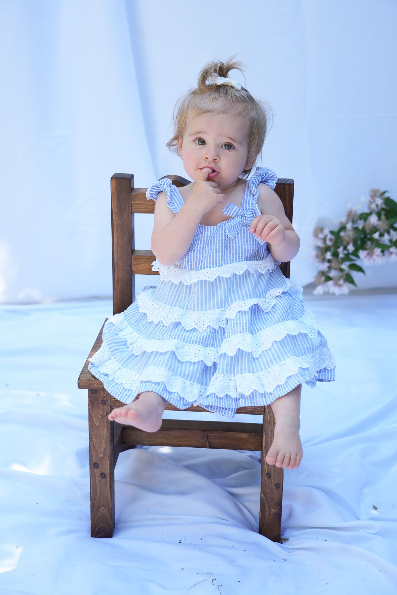 Adorable blonde toddler girl in blue ruffled dress sitting on small wooden chair with finger in mouth.