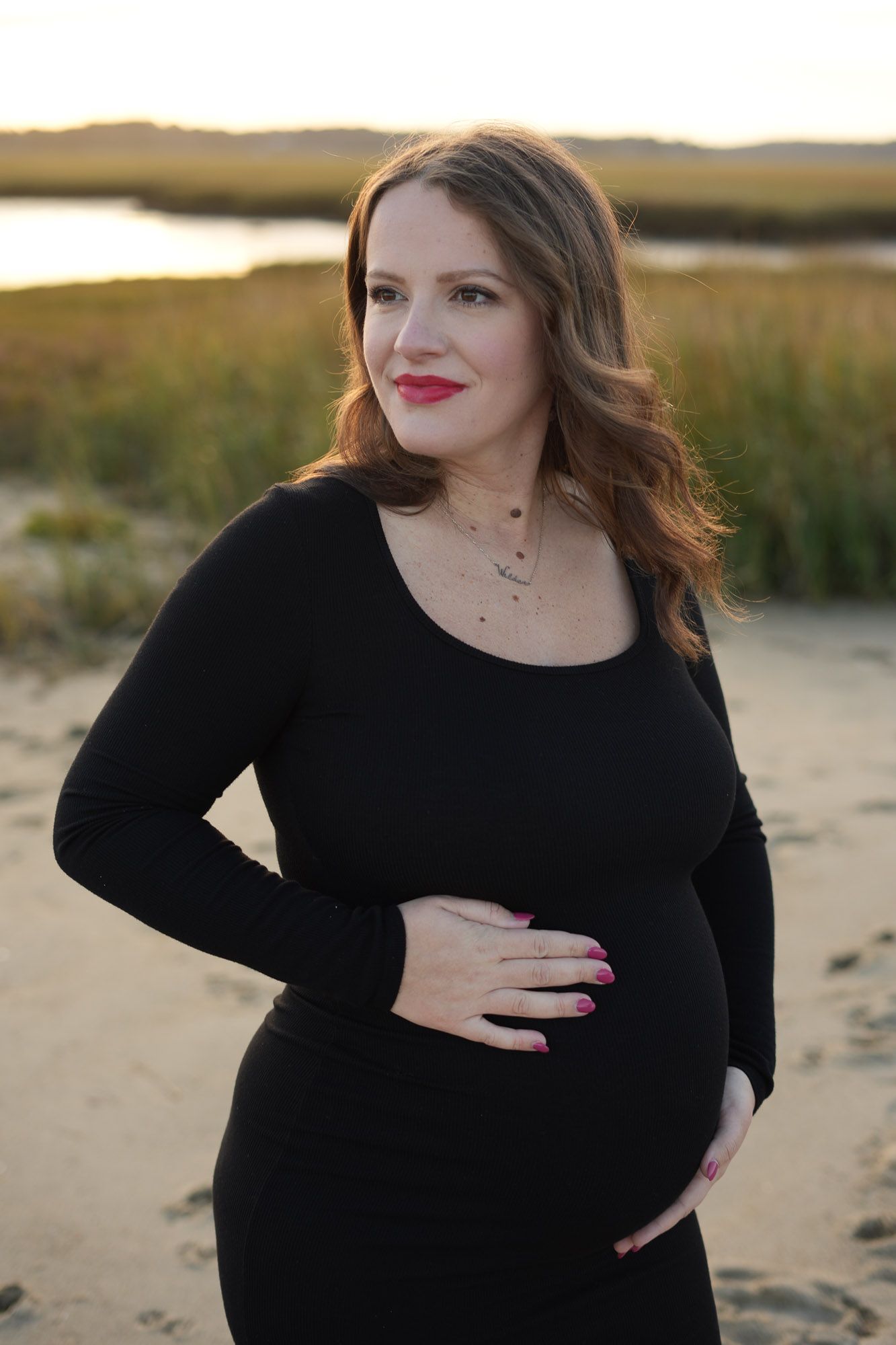 Smiling pregnant woman in black dress holding belly on beach at golden hour.