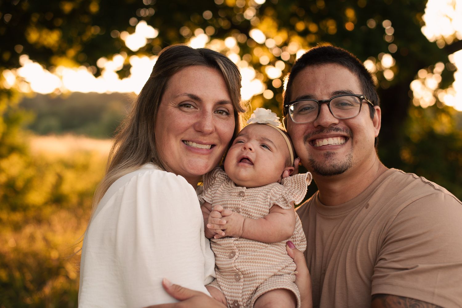 Happy family of three smiles together outdoors, parents holding their laughing baby in golden light.