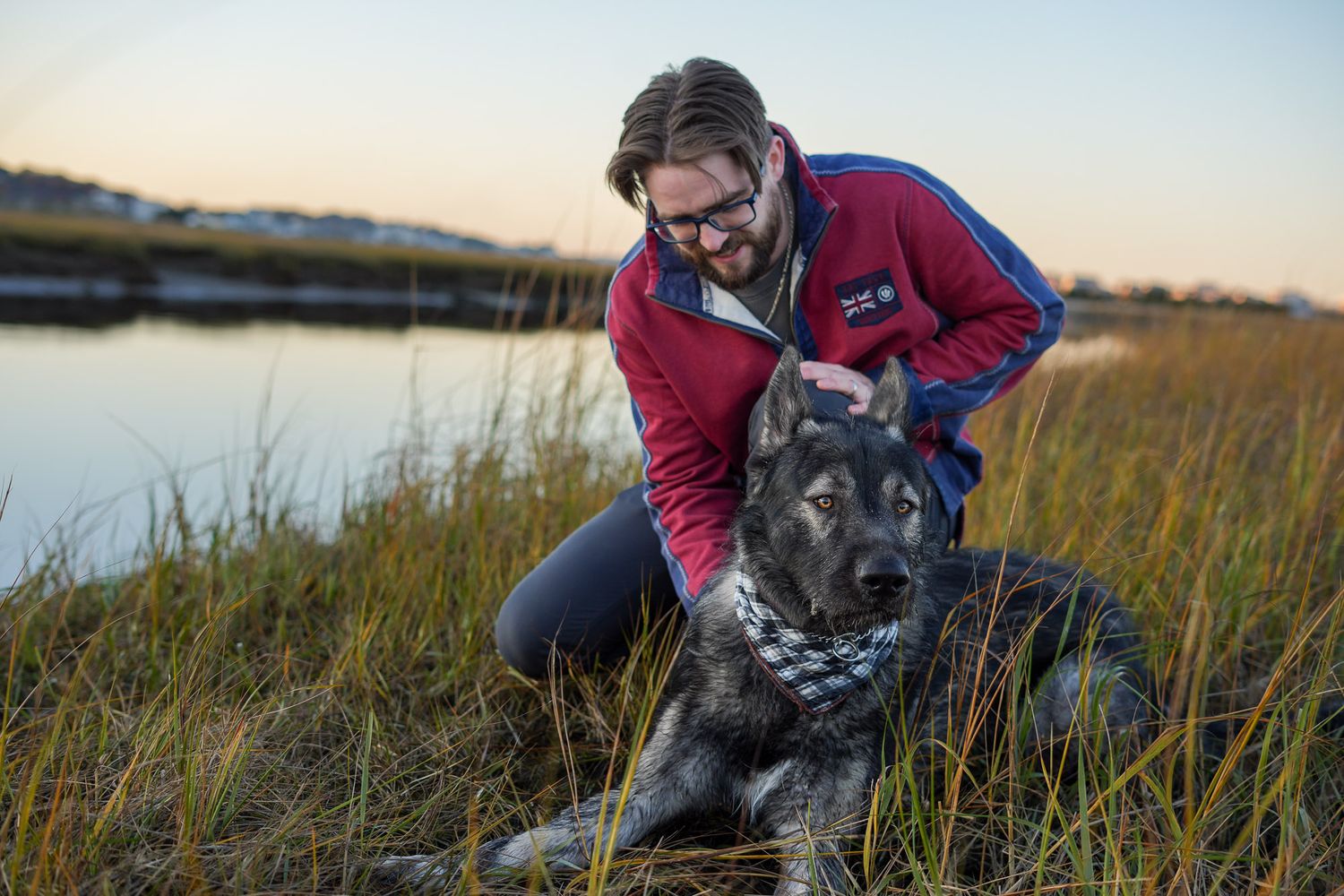 Man with glasses petting a gray German Shepherd dog in tall grass near a calm waterway at sunset.
