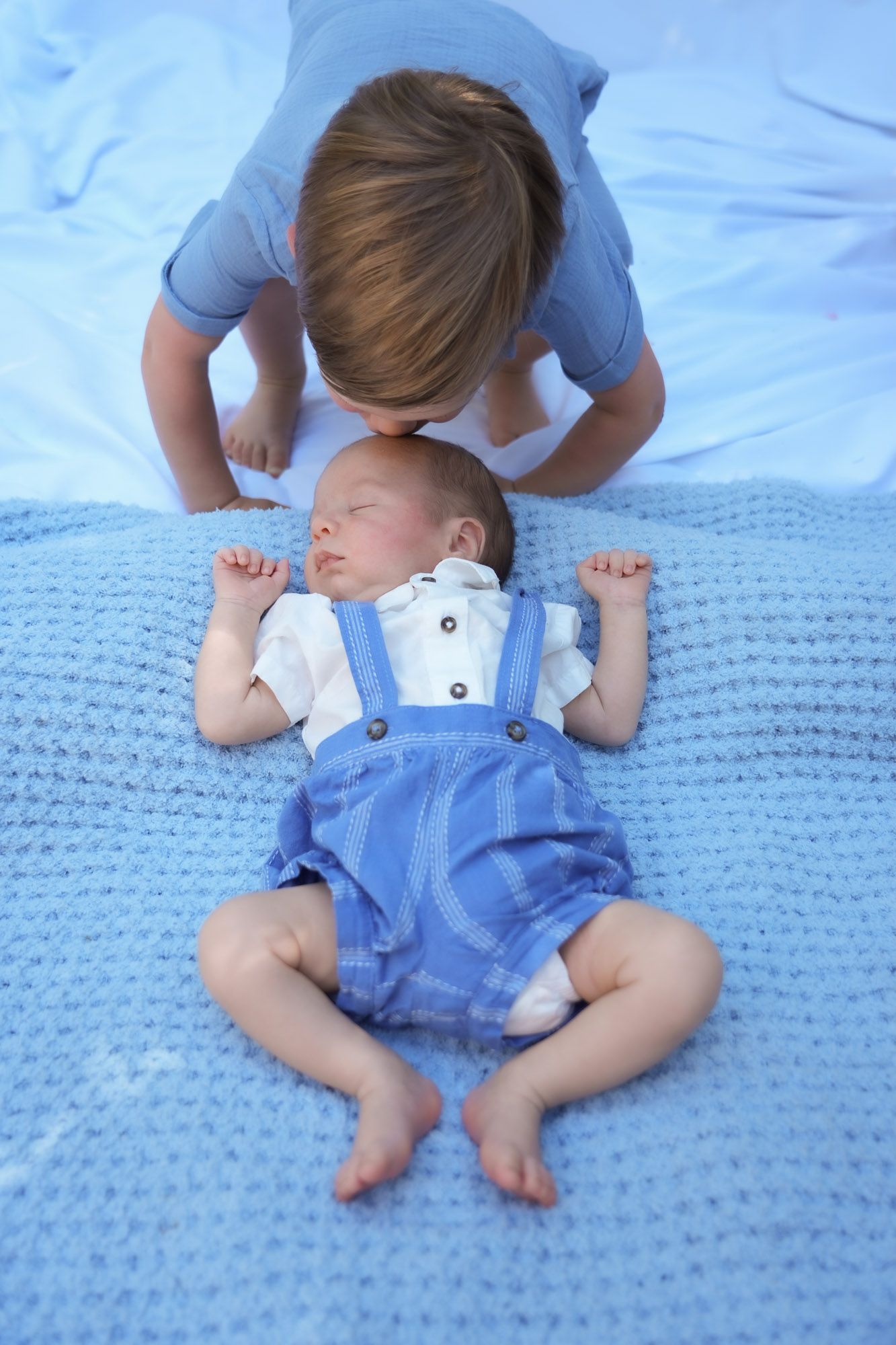 Young child gently kissing sleeping newborn baby lying on blue knit blanket in matching outfits.