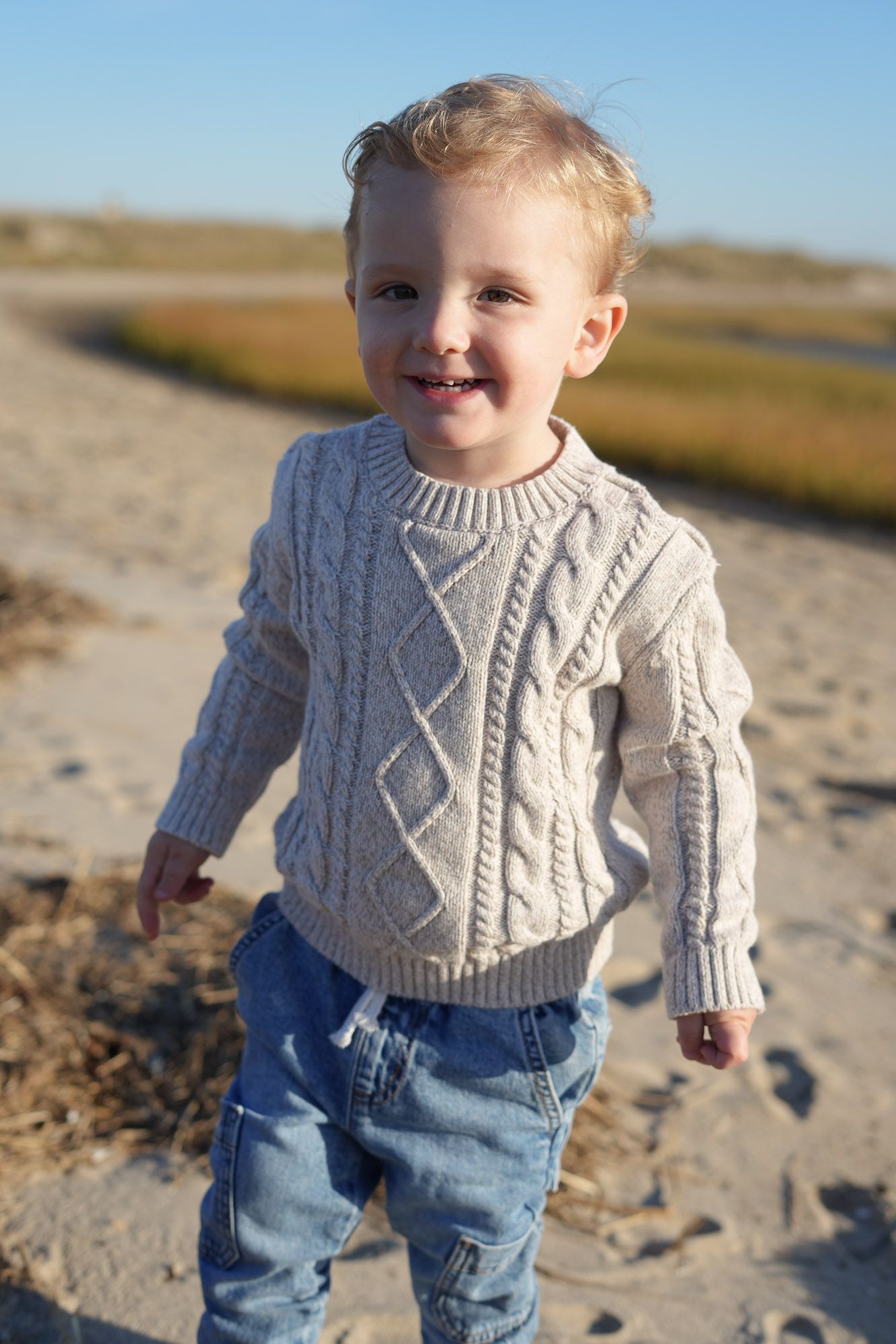 Happy toddler boy smiling on sandy beach wearing cream cable-knit sweater and jeans on sunny day.