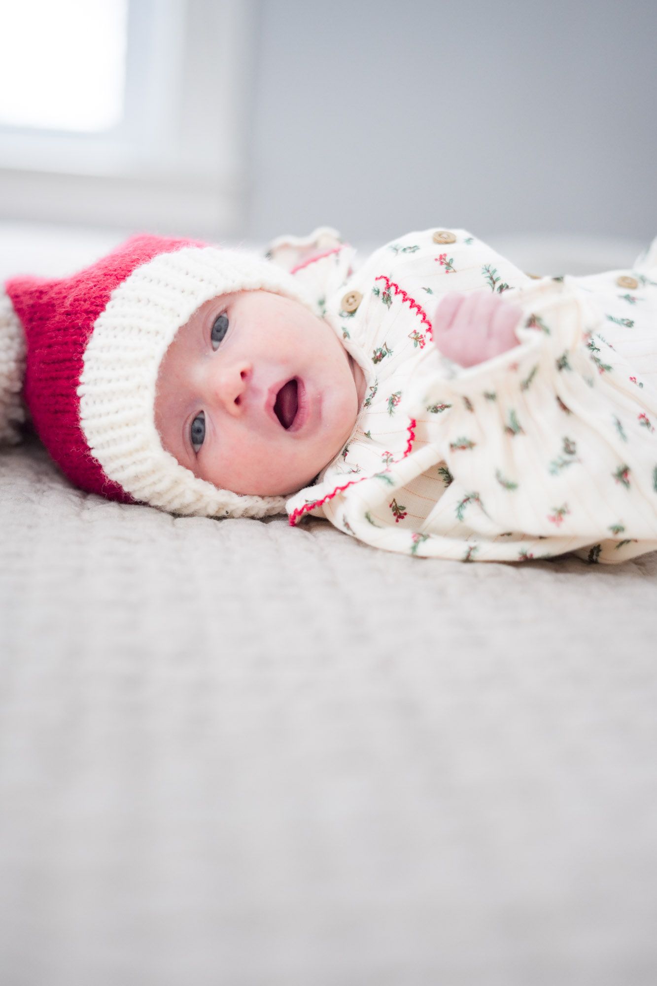Adorable newborn baby wearing red and white knit Santa hat lying on a soft gray blanket indoors.