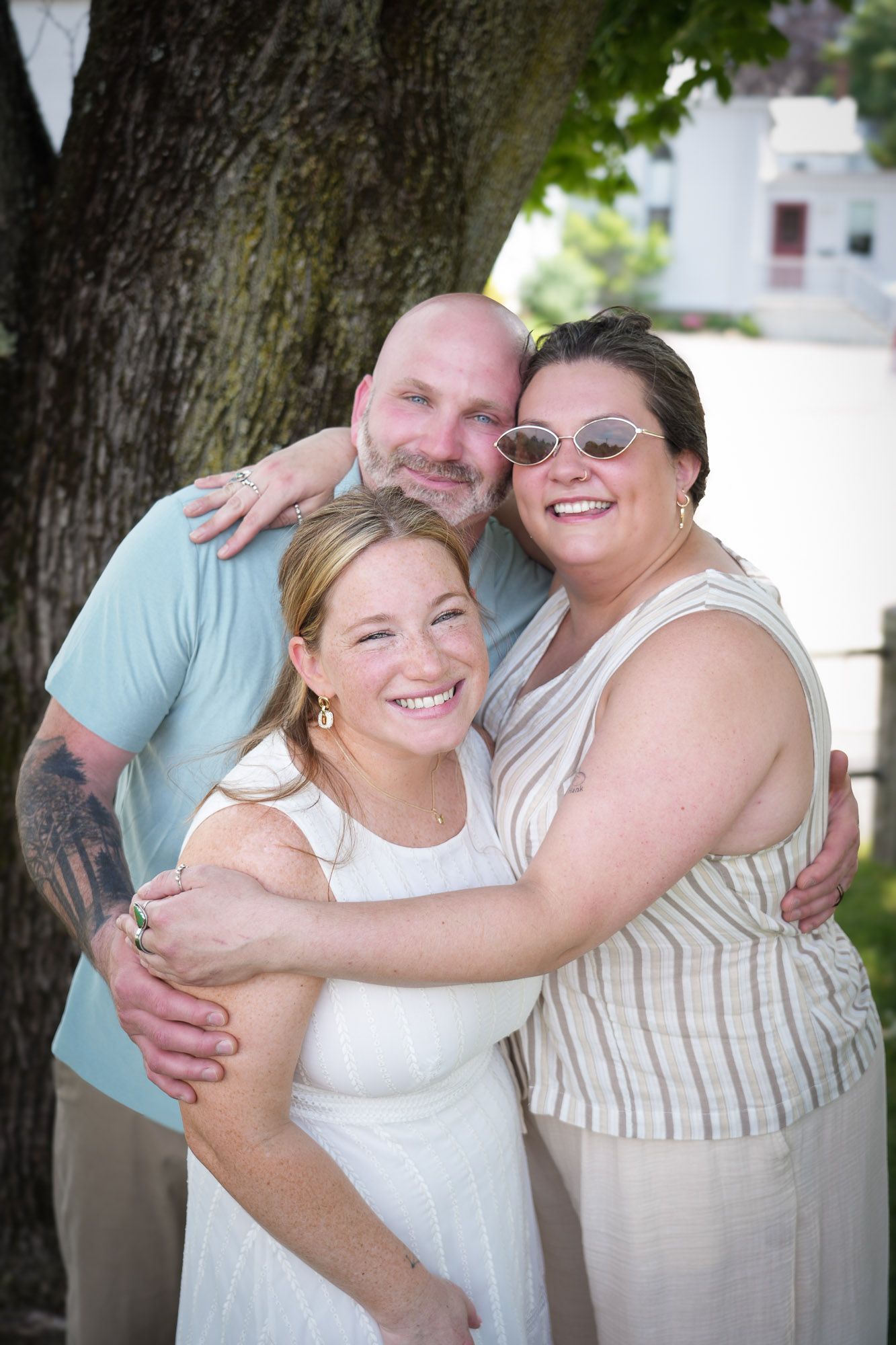 Three people smiling and hugging outdoors near a large tree, celebrating a special occasion.