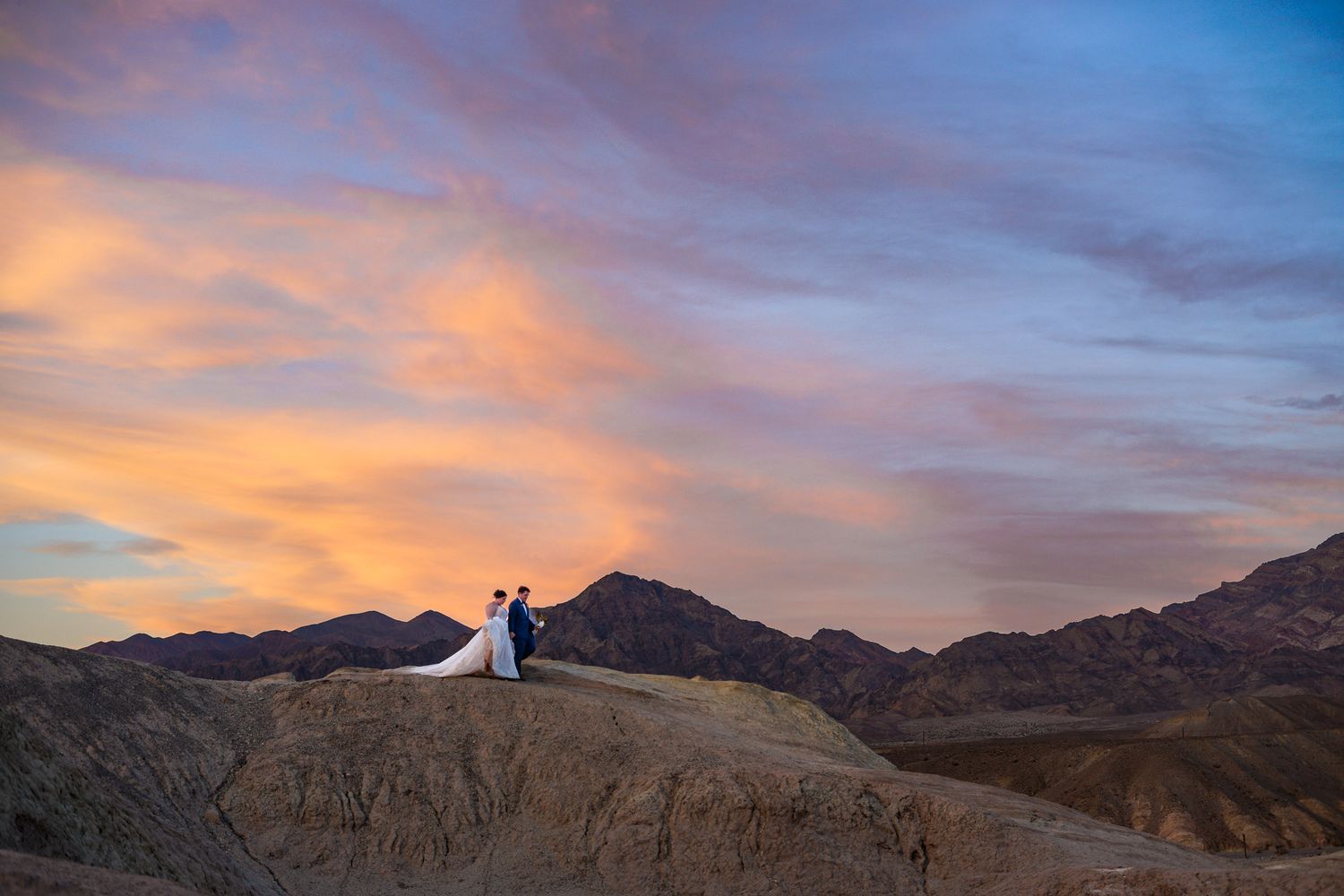 Bride and groom stand on desert rock formation beneath dramatic colorful sunset sky in Death Valley National Park.