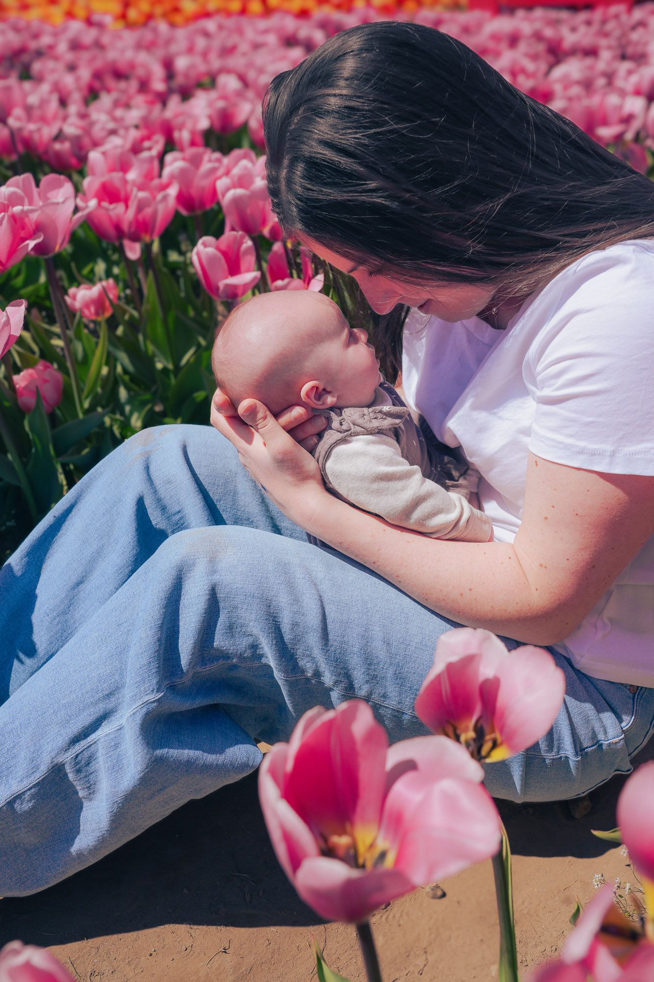 A mother tenderly holds her sleeping newborn baby while sitting among rows of pink tulips.