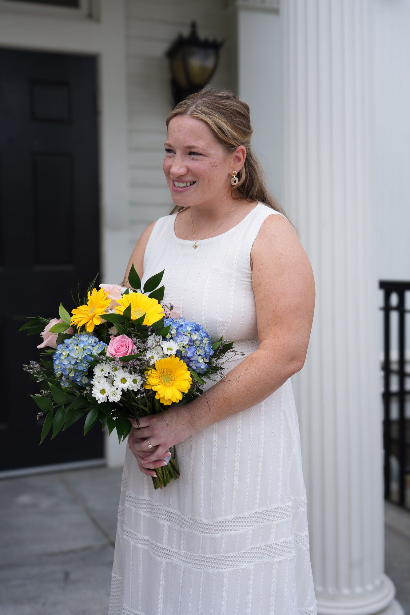 Smiling bride in white sleeveless dress holding colorful bouquet of yellow, blue, and pink flowers on porch.