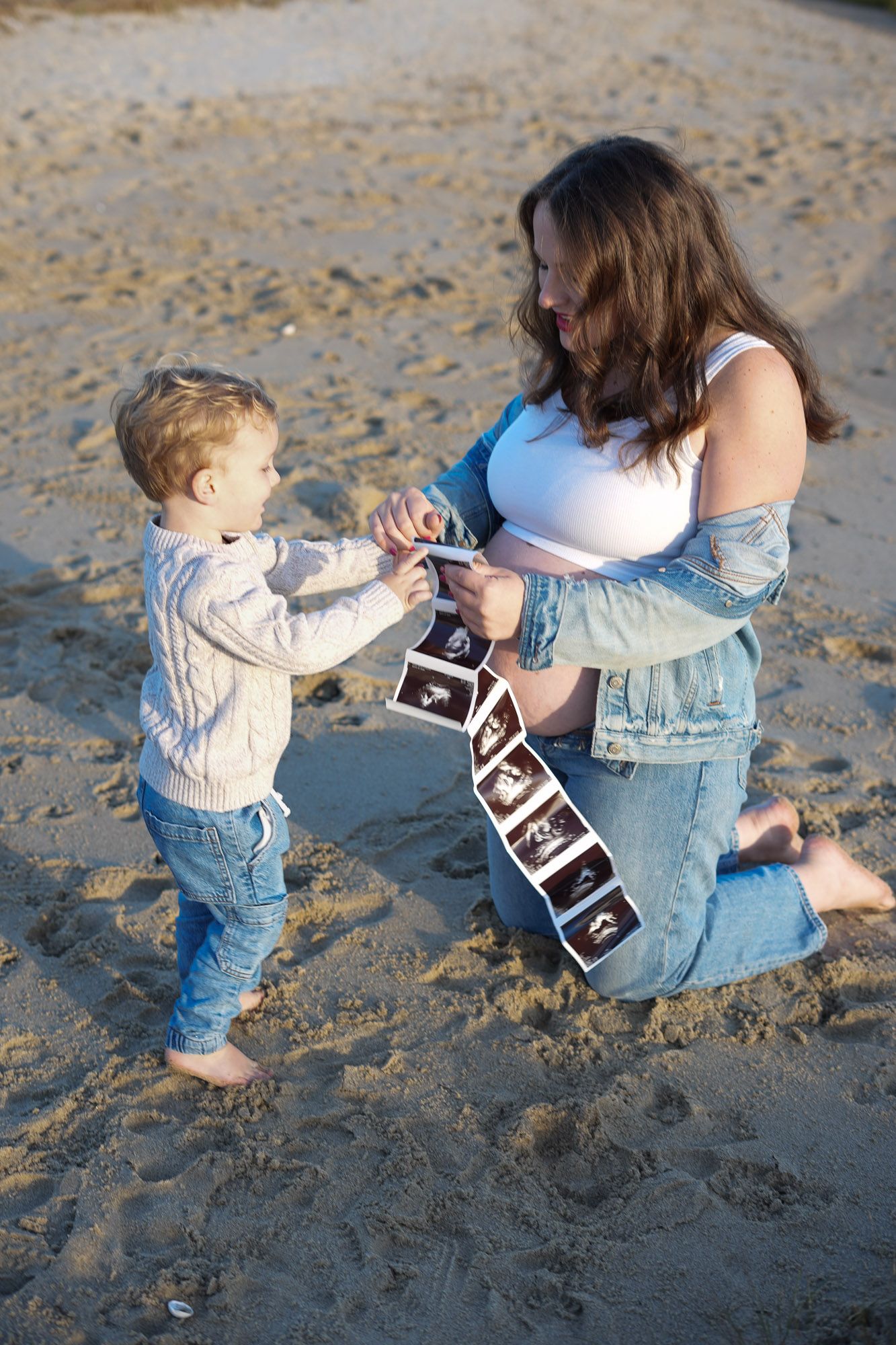 Pregnant woman kneeling on beach showing ultrasound photos to her young toddler son on sandy shore.