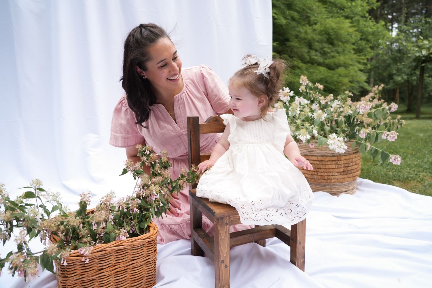 Smiling mother in pink dress kneels beside toddler daughter in white dress on wooden chair surrounded by flower baskets.
