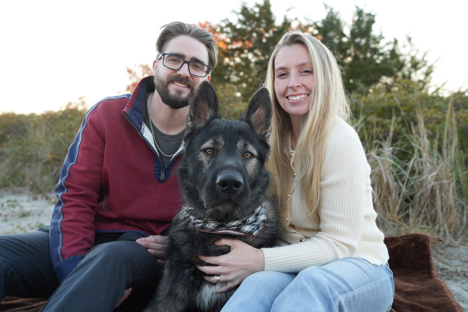 Smiling couple posing with their young German Shepherd dog on a sandy beach at sunset.