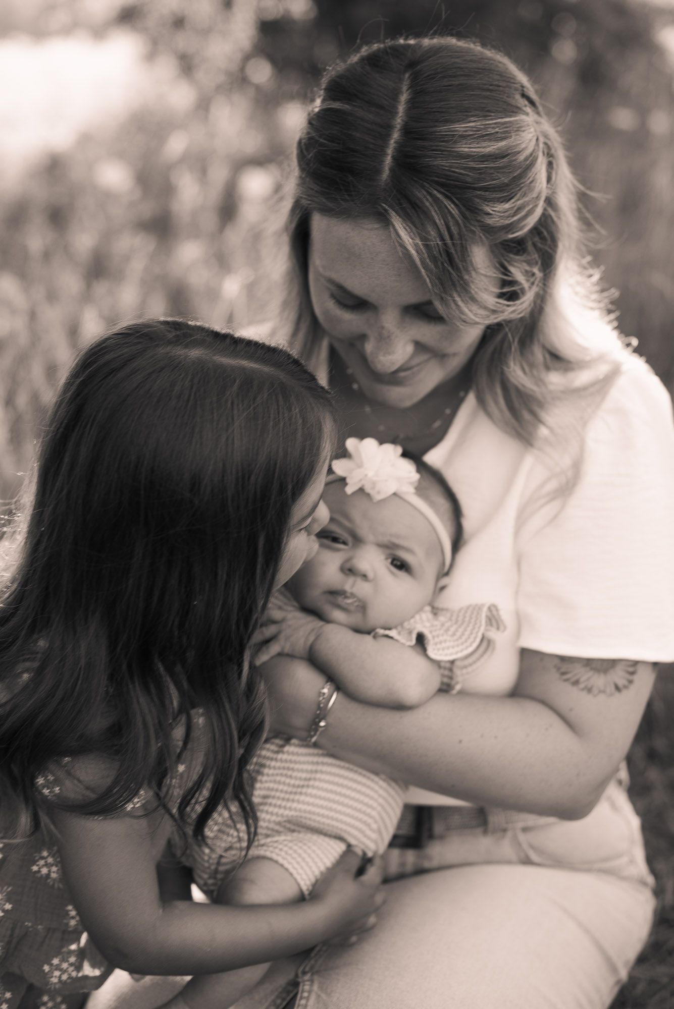 Black and white photo of mother smiling at newborn baby held by older sister.
