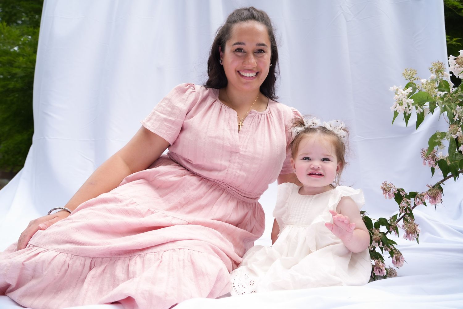Smiling mother in pink dress sits beside cheerful toddler daughter in white dress against white fabric backdrop outdoors.