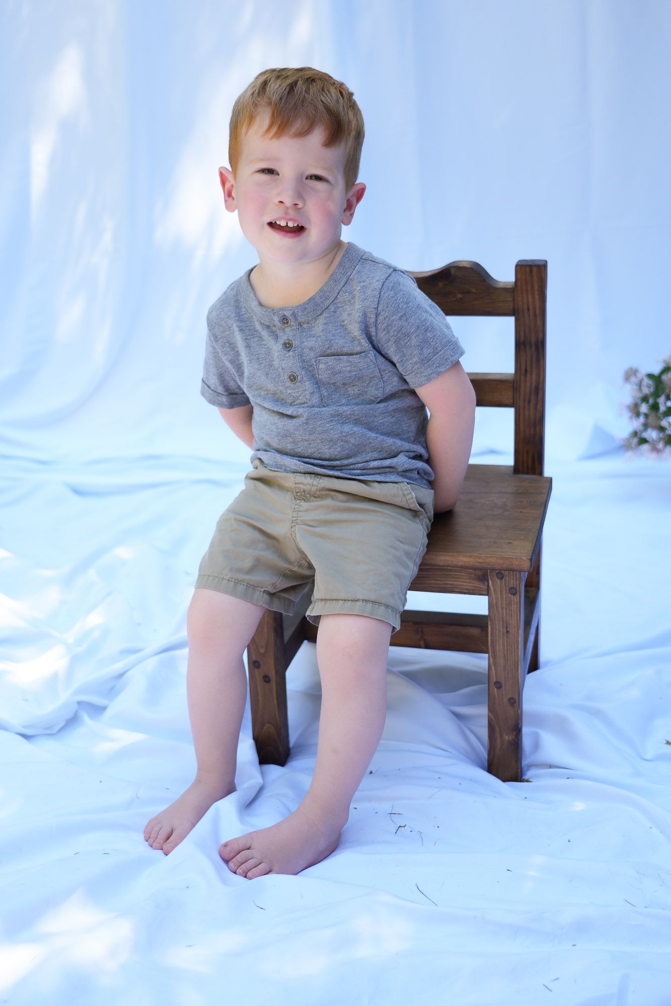 Happy redhead boy laughing while seated on a small wooden chair on white fabric.