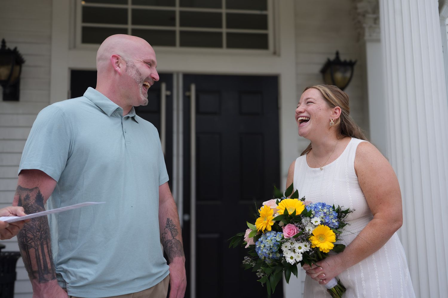 Bride and groom laughing together on white columned porch, bride holding vibrant yellow and blue flower bouquet.