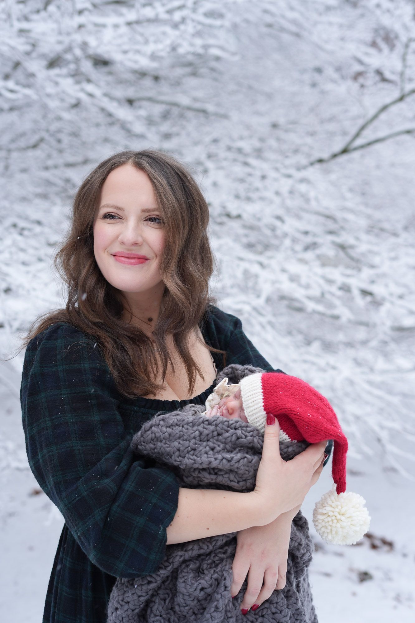 Smiling mother holding swaddled newborn wearing red Santa hat in snowy winter outdoor setting.