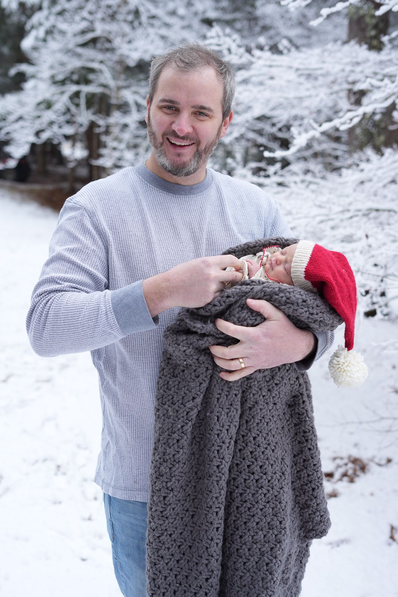Proud smiling father holding swaddled newborn in red Santa hat in snowy winter setting.