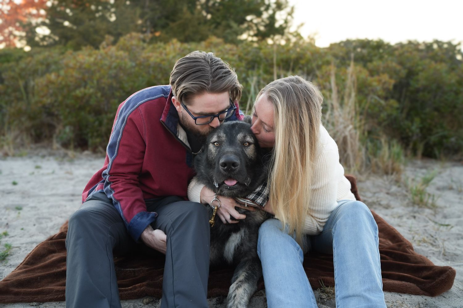 Couple leaning in to kiss their happy German Shepherd dog on a sandy beach blanket.