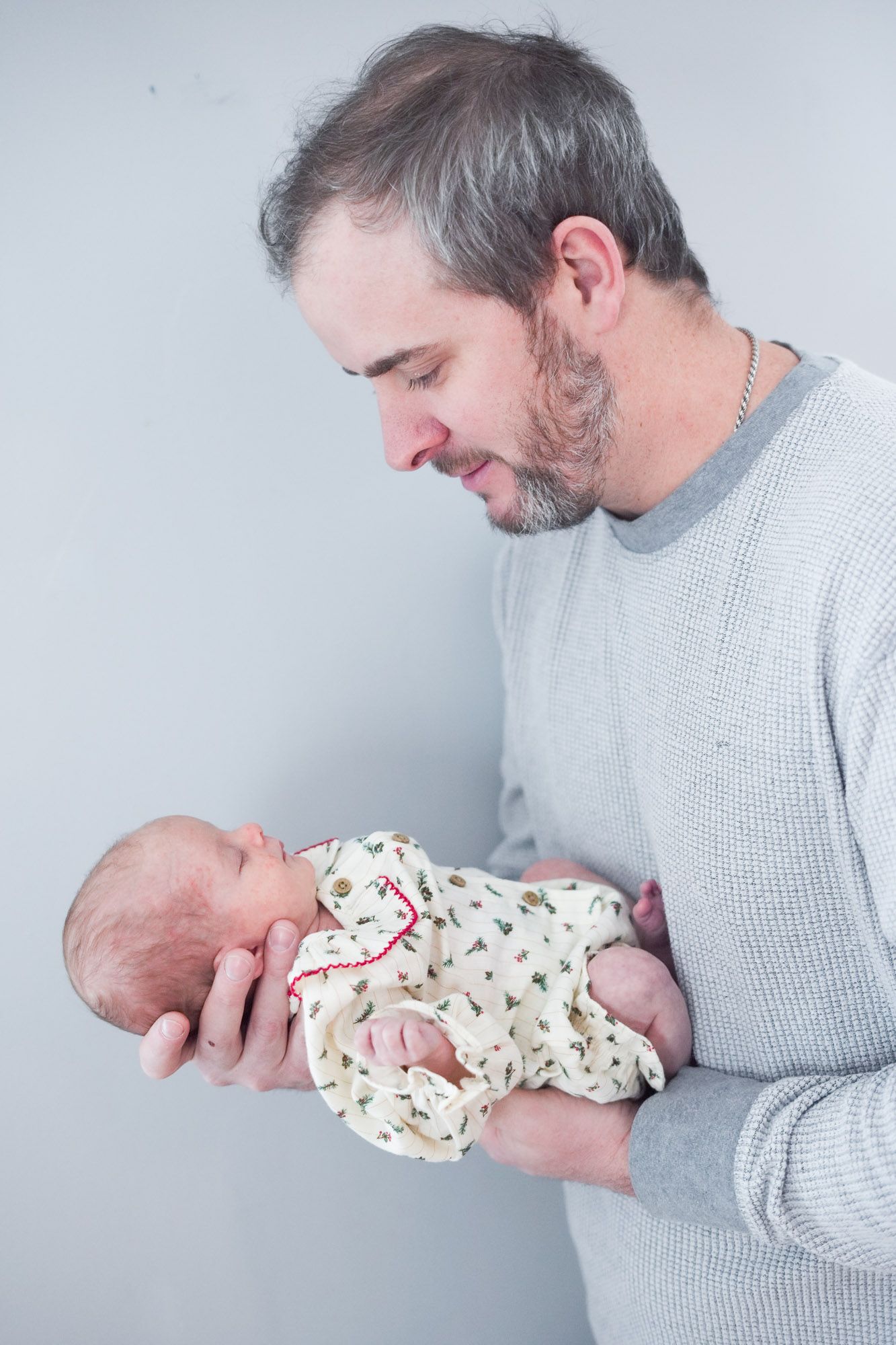 Father in gray sweater gently cradling his sleeping newborn baby in both hands.