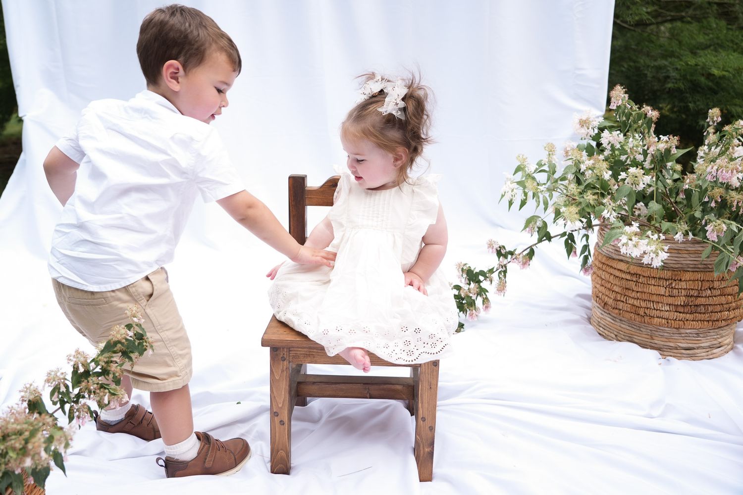 Young boy leans toward laughing toddler girl in white lace dress on wooden chair with pink flower arrangements nearby.