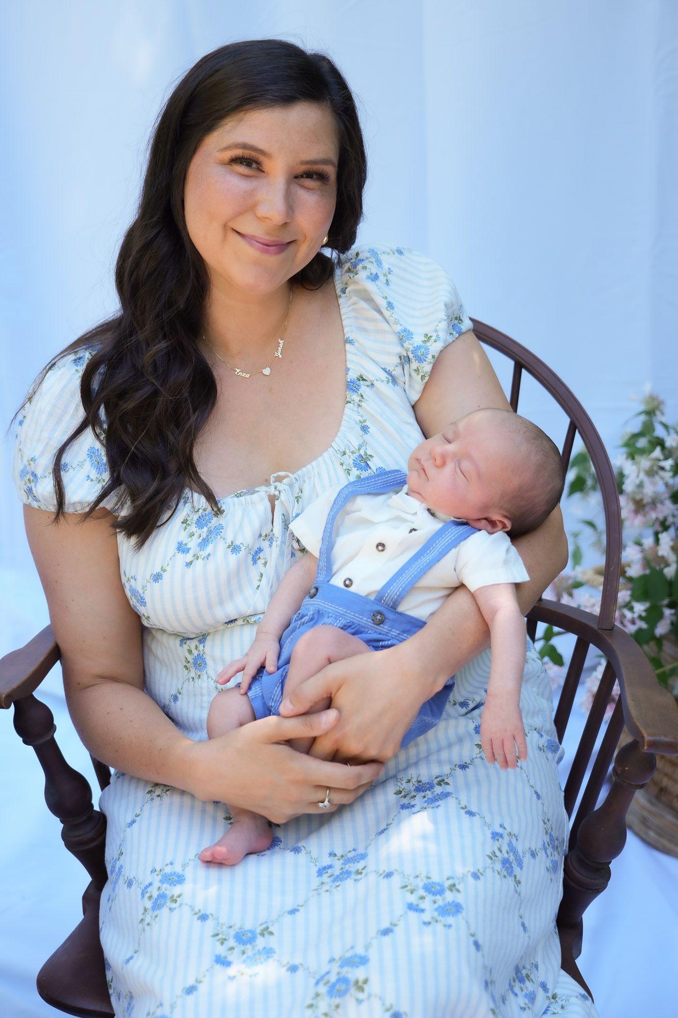 Mother in floral dress holding sleeping newborn baby in blue overalls.