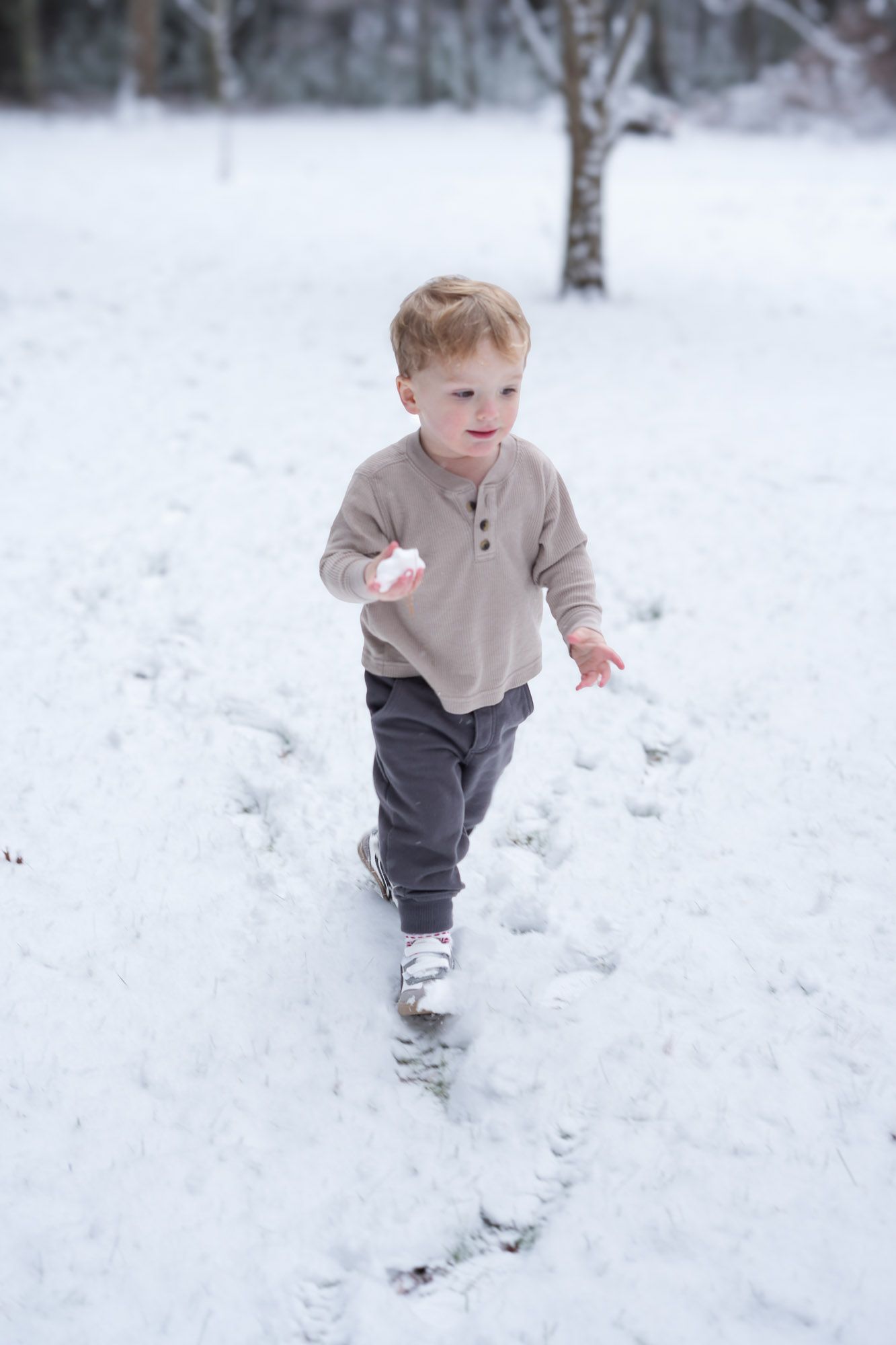 A young toddler boy with red hair plays in the snow, holding a snowball while walking through a winter wonderland.