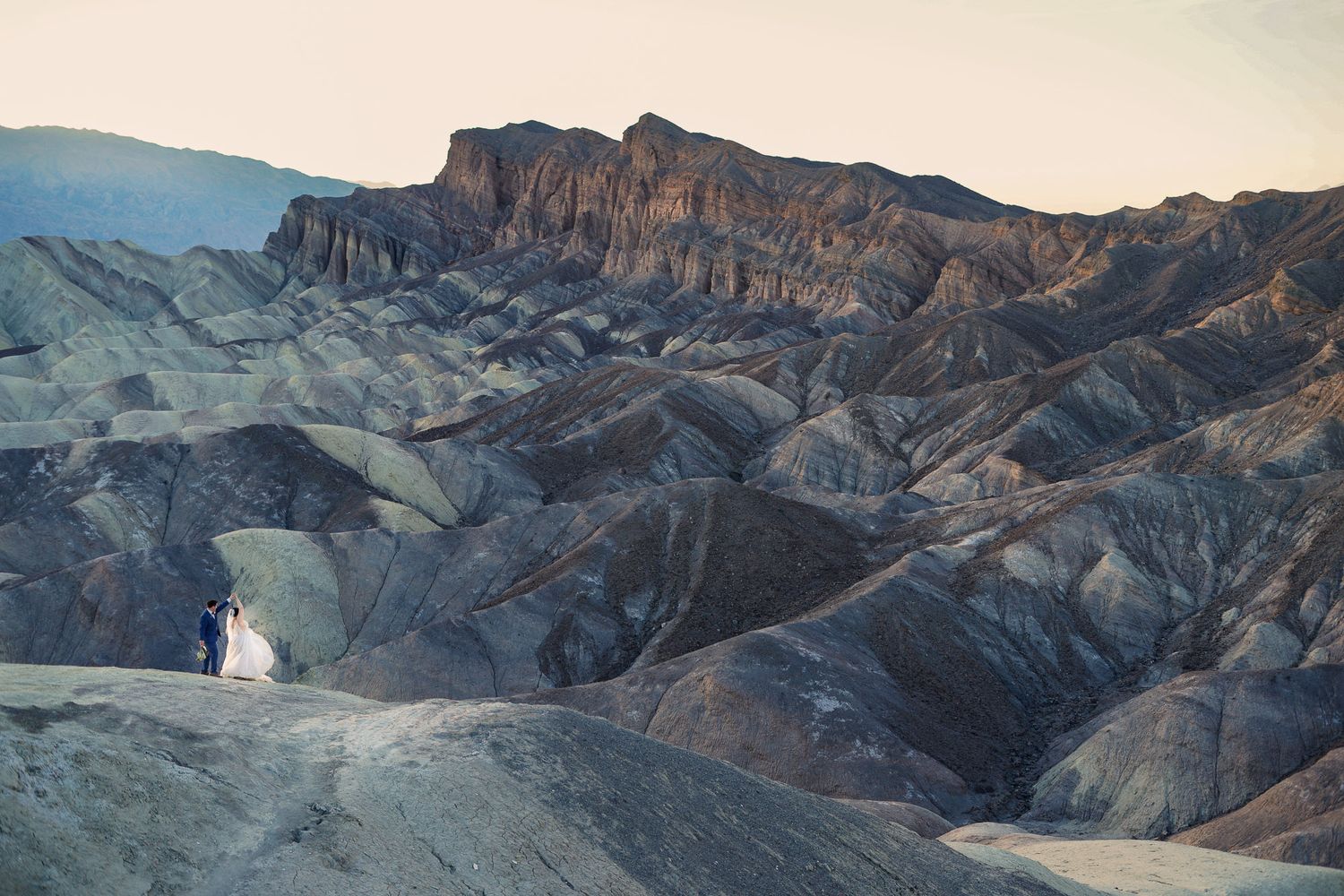 Bride and groom appear small against dramatic eroded hills of Death Valley National Park at sunset.
