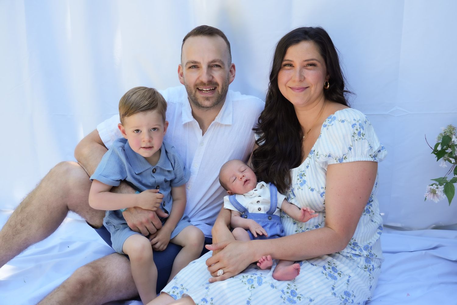 Family portrait with parents, toddler son, and newborn baby on white fabric backdrop.