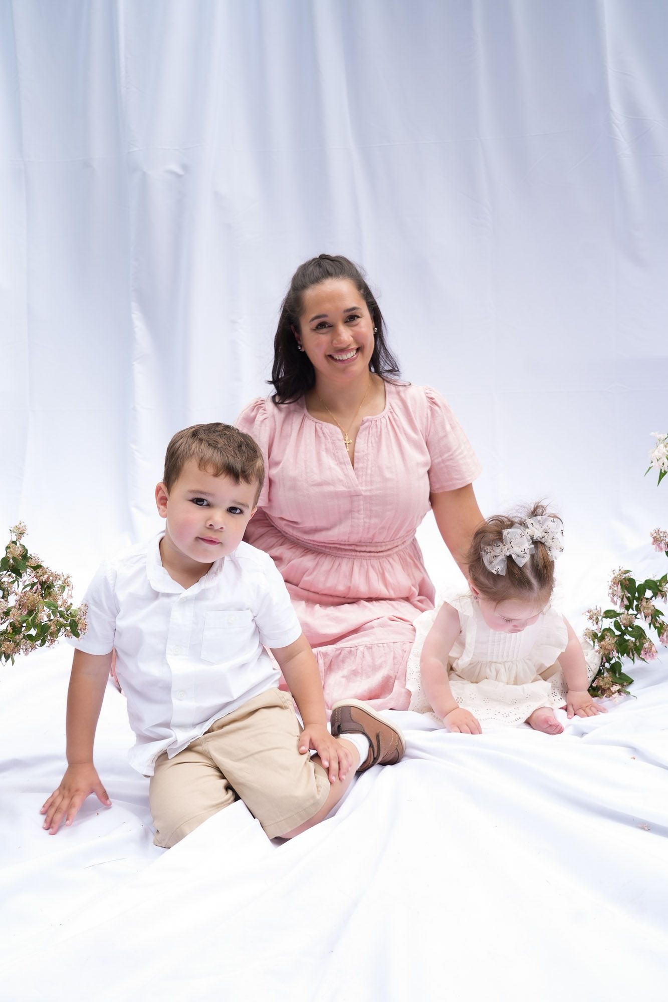 Mother in pink dress sitting with toddler boy and baby girl on white backdrop at photo session.