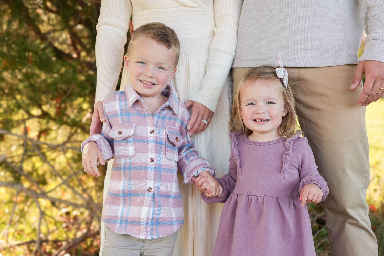 Young boy and girl smiling and holding hands with parents in front of autumn foliage outdoors.