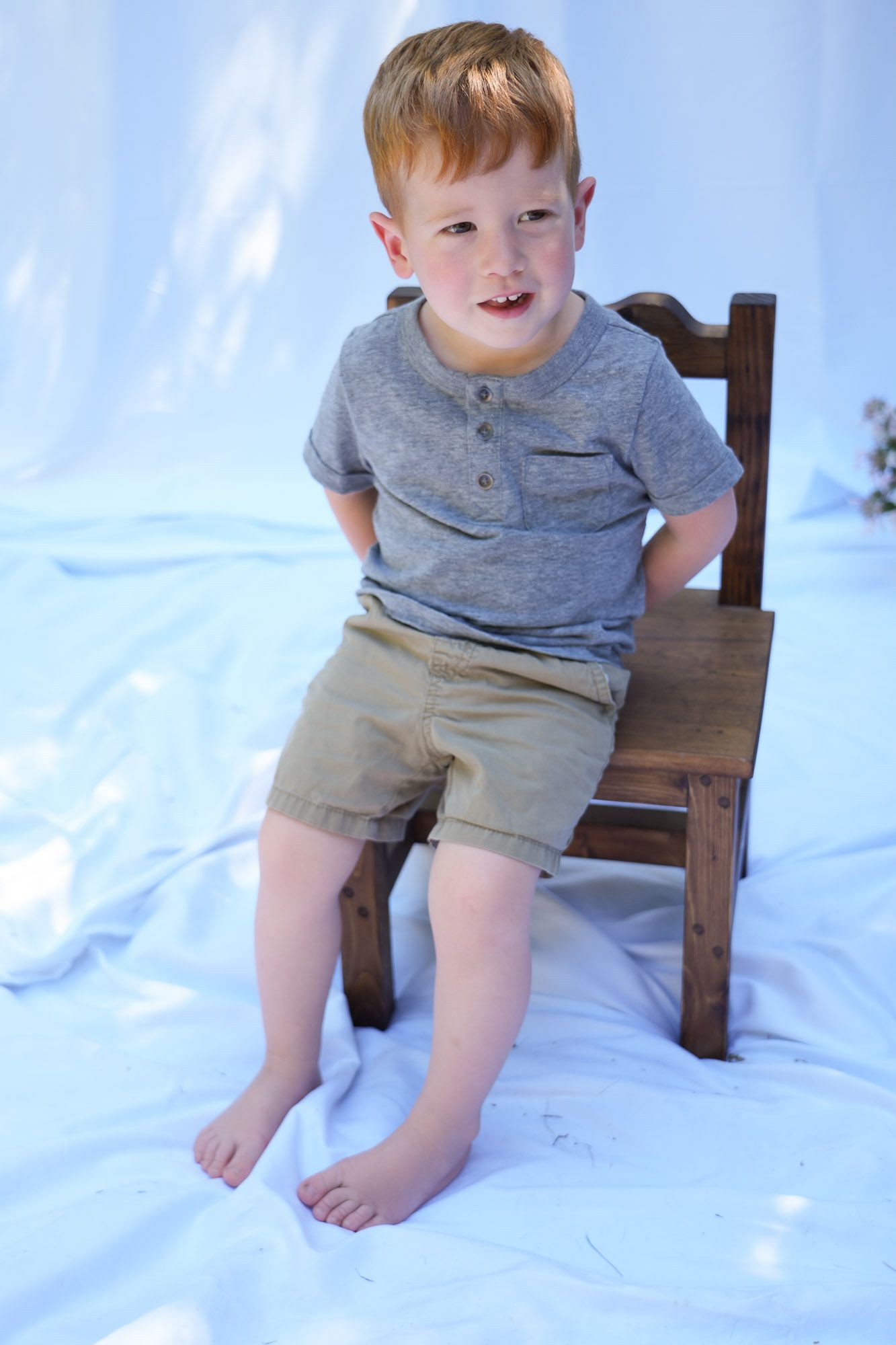 Young red-haired boy in gray shirt and khaki shorts sitting on small wooden chair.
