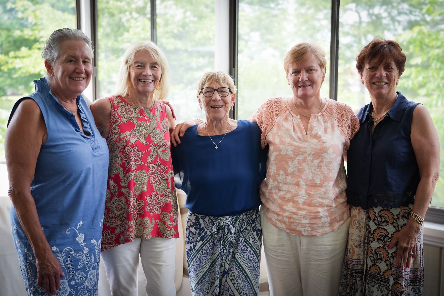 Five smiling women posing together indoors near large windows with green trees visible in the background.