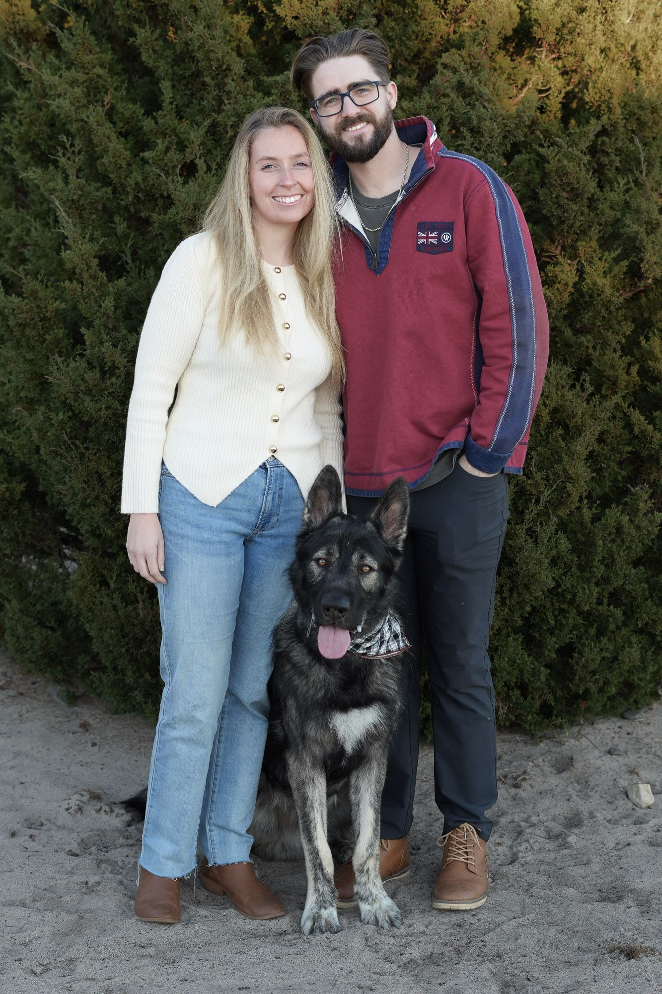 Smiling couple in casual fall outfits posing with their German Shepherd dog on sandy ground near evergreens.