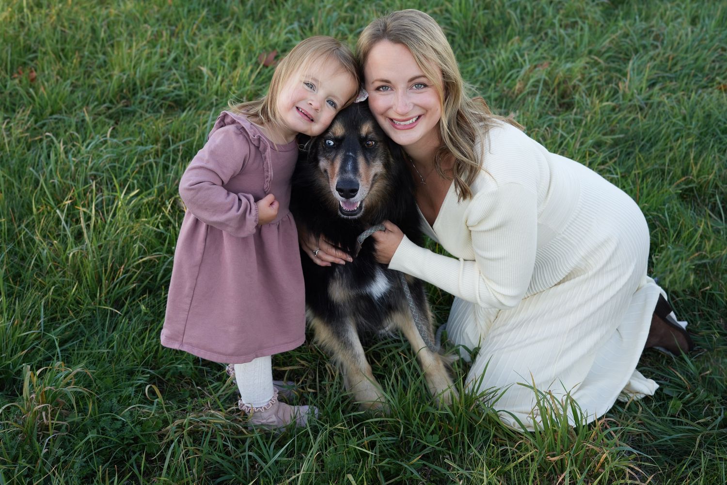 A smiling woman and young girl pose with their black and brown dog on grass.