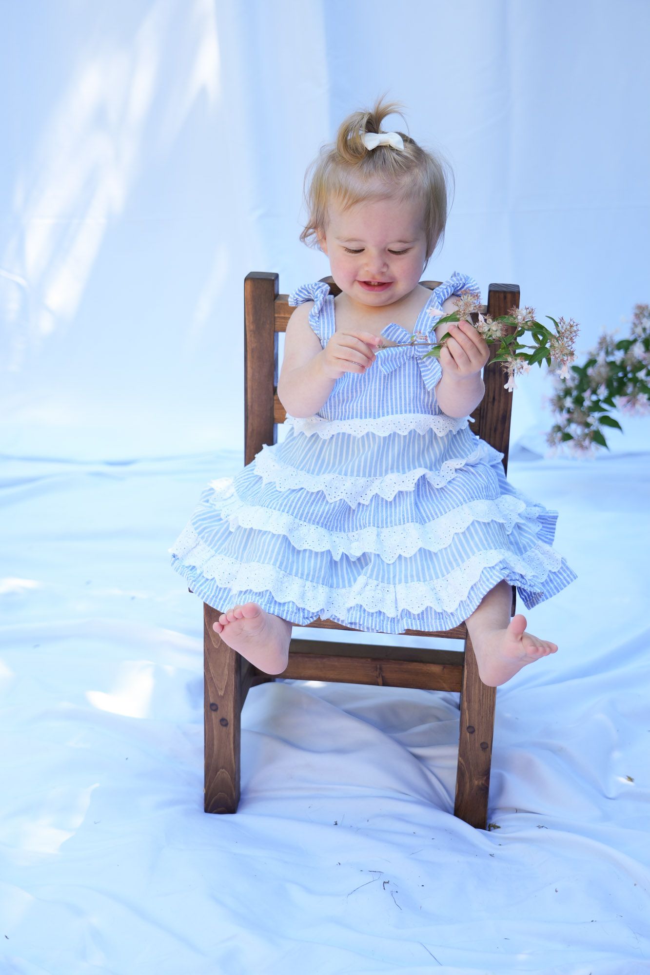 Cute baby girl in blue striped ruffle dress sitting on wooden chair examining small flowers.