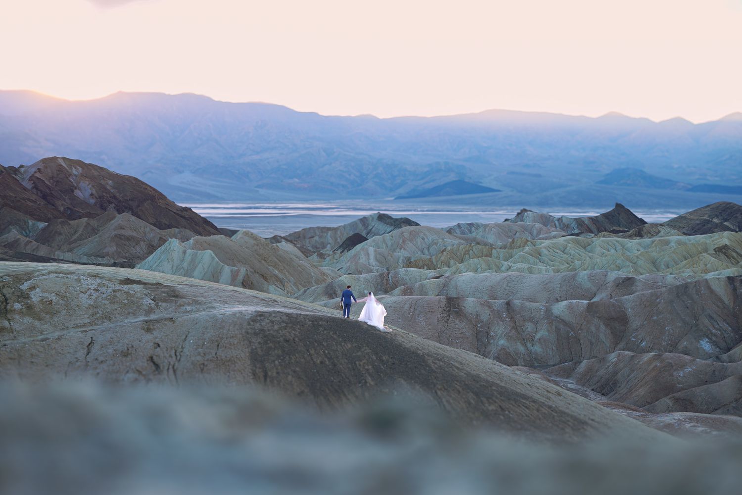 Bride and groom standing amid stunning Death Valley badlands landscape at sunset with distant mountains.