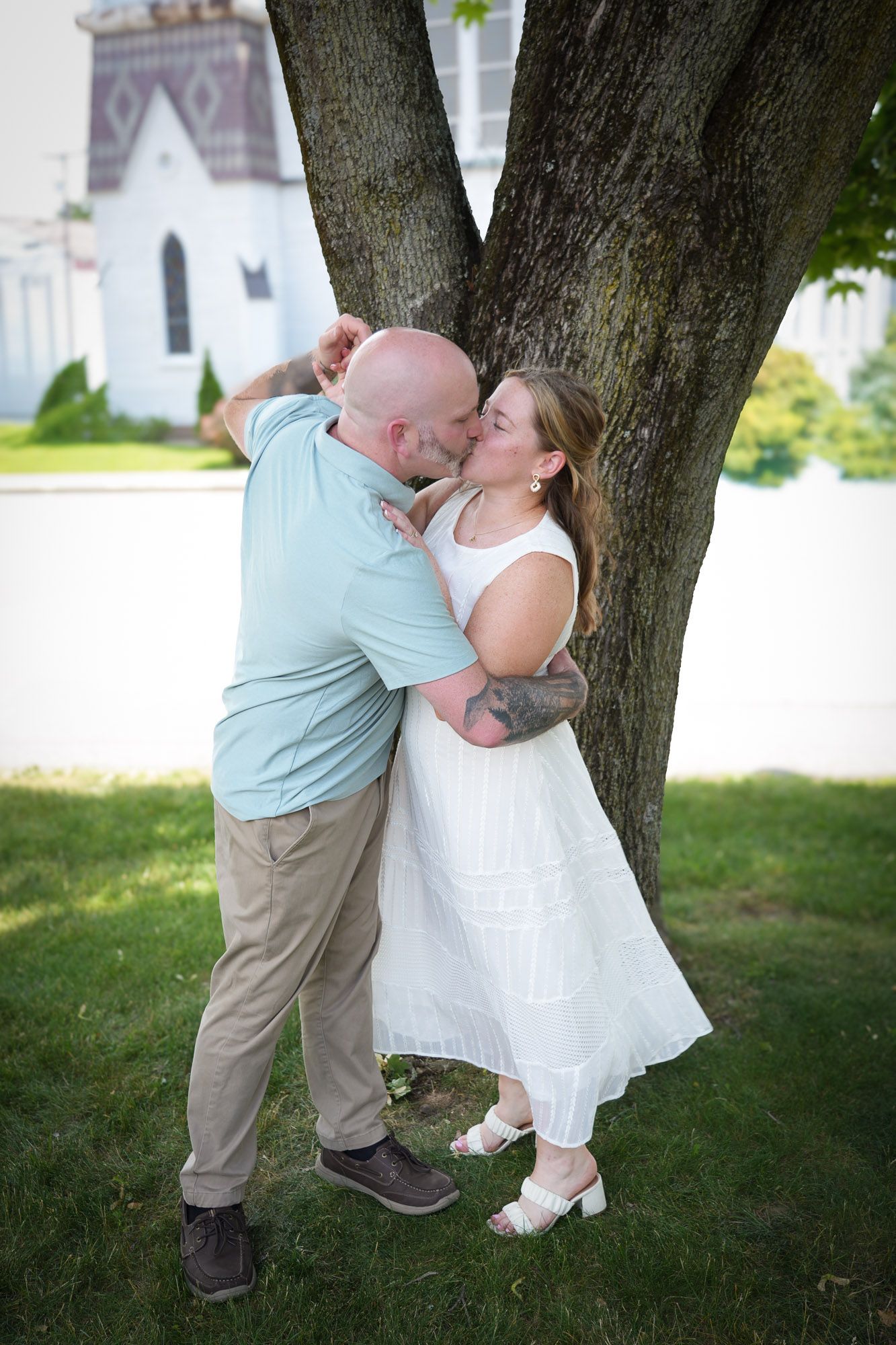 A couple shares a romantic kiss leaning against a large tree with a white church in the background.
