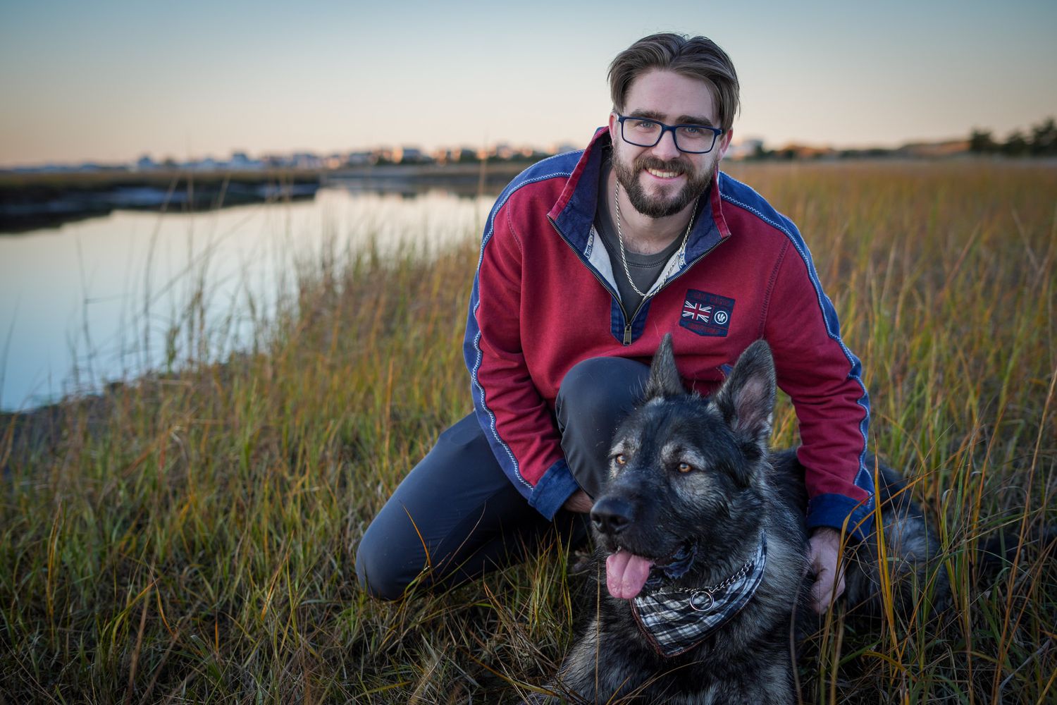 Smiling man in red jacket crouching beside his German Shepherd dog in a marsh field at sunset.