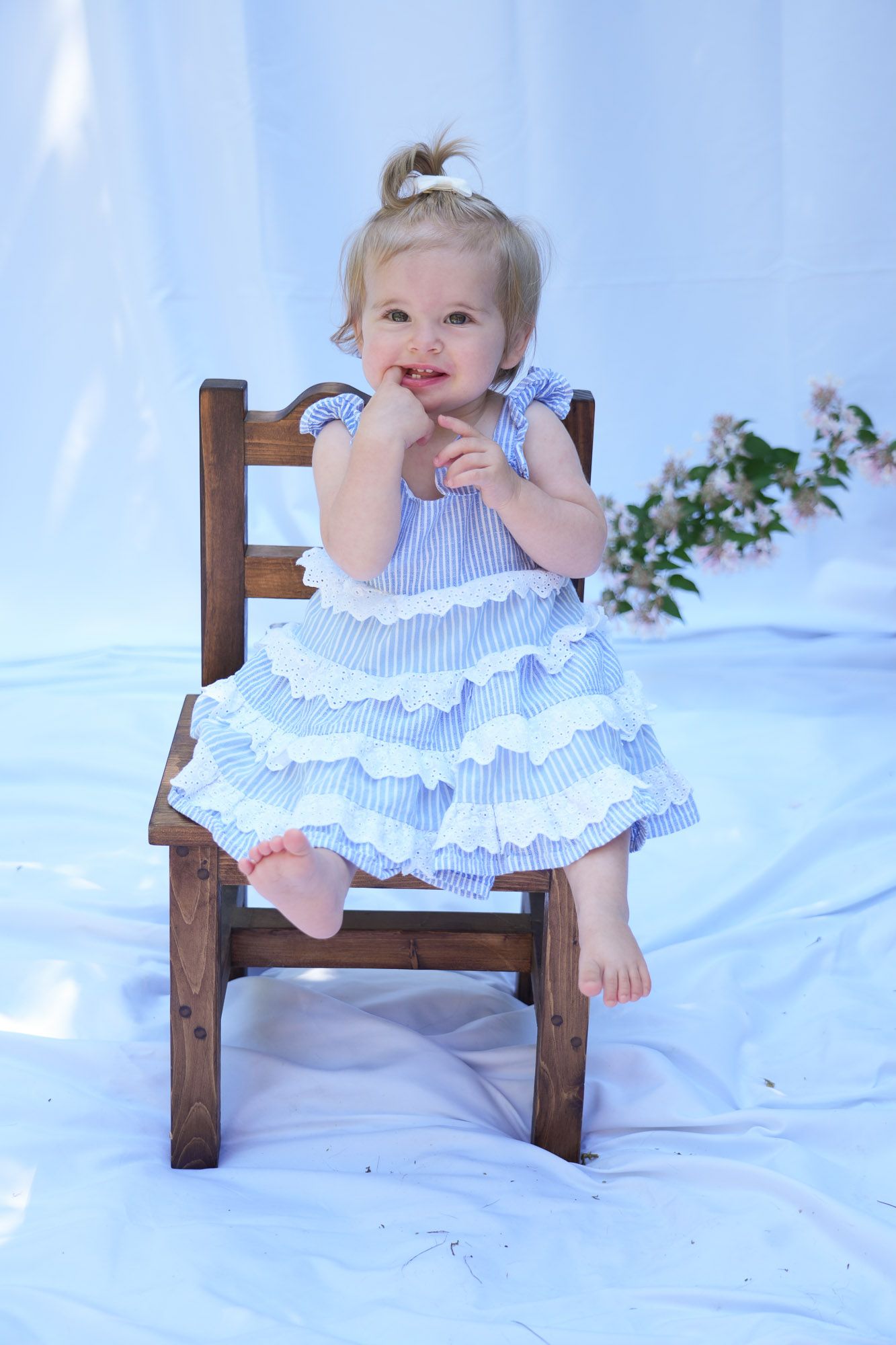 Cute blonde toddler girl in blue striped ruffled dress smiling while seated on a small wooden chair.