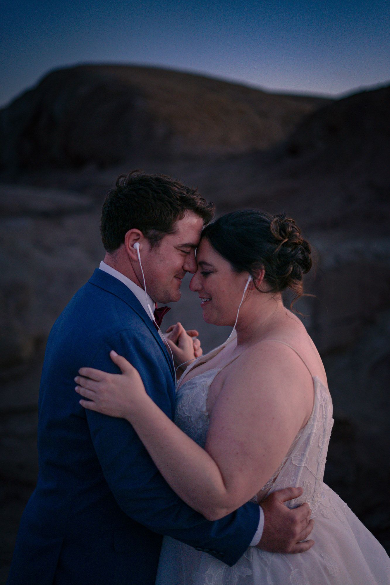 Couple sharing an intimate moment in wedding attire at dusk in a rocky desert landscape, sharing earbuds.